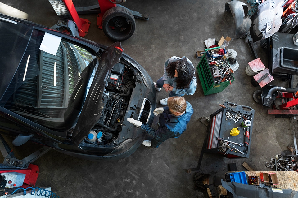 Overhead view of a mechanic and a customer standing beside a car with its hood open inside an auto repair shop. Tools, toolboxes, and various equipment are scattered around the concrete floor as they inspect the vehicle’s engine bay.