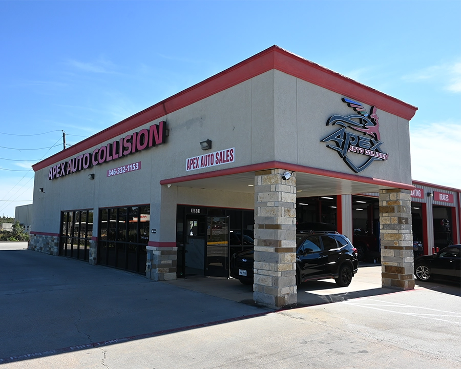 Exterior view of the Apex Auto Collision building, featuring a large metal logo with a stylized car and wings mounted on the front facade. The entrance area has stone pillars, a shaded walkway, and a glass door, with garage bay doors visible to the right. A sign for Apex Auto Sales is displayed on the corner of the building.