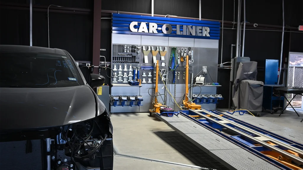 A vehicle frame straightening area inside an auto body shop, featuring a CAR-O-LINER system with a large blue-and-silver tool wall mounted behind it. The wall is organized with clamps, brackets, wrenches, and alignment tools. The frame machine platform extends into the foreground, while the partially disassembled front end of a dark-colored car is visible on the left side.