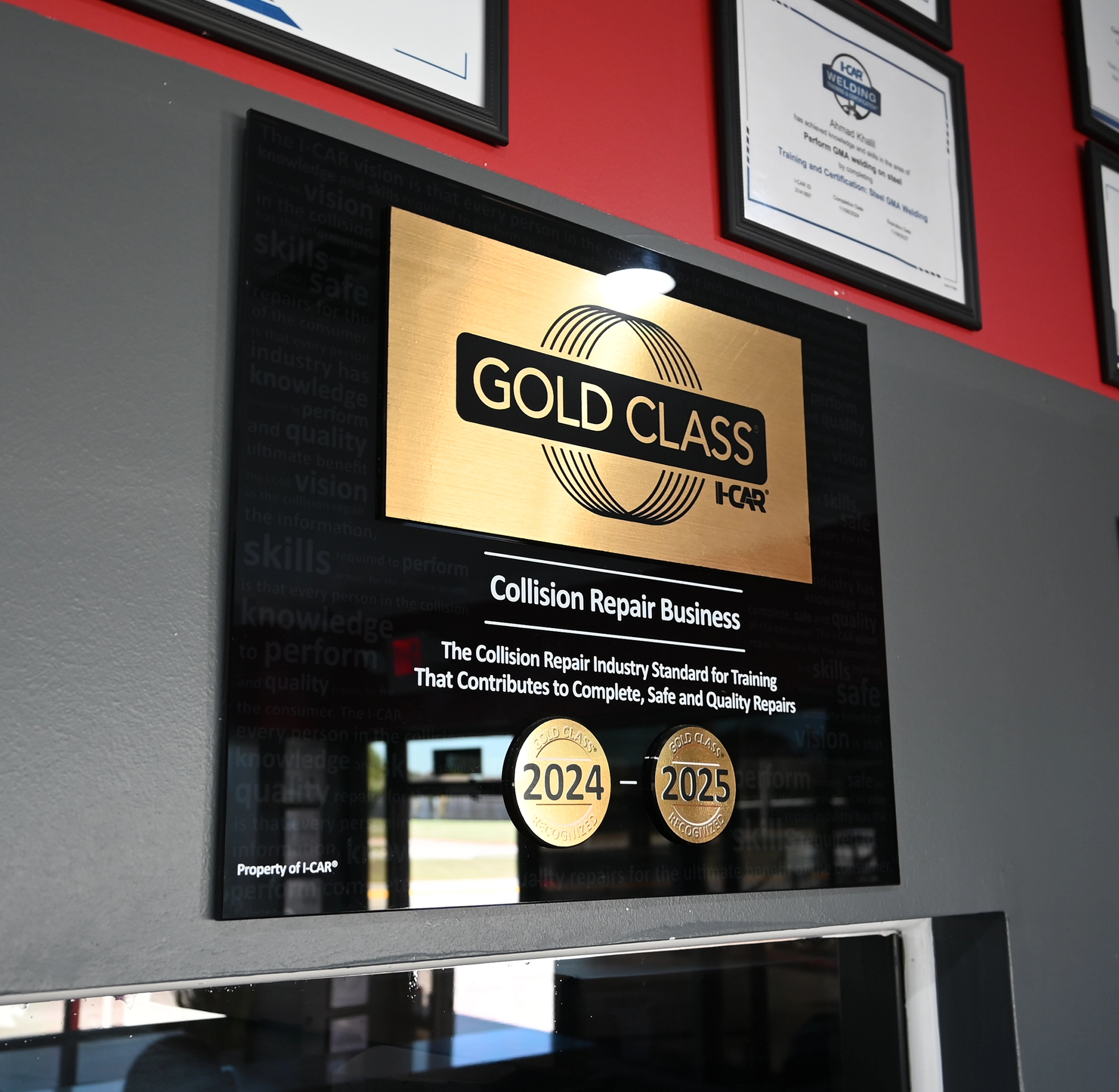 A wall display featuring an I-CAR Gold Class plaque for a collision repair business, showing certification years 2024–2025. Surrounding the plaque are multiple framed training certificates and a RepairPal Certified sign, all mounted on a red and gray interior wall.
