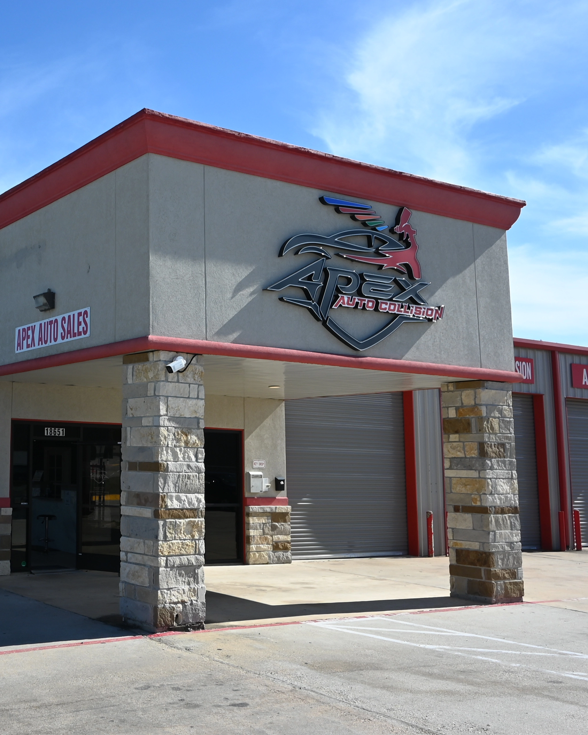 Exterior view of the Apex Auto Collision building, featuring a large metal logo with a stylized car and wings mounted on the front facade. The entrance area has stone pillars, a shaded walkway, and a glass door, with garage bay doors visible to the right. A sign for Apex Auto Sales is displayed on the corner of the building.