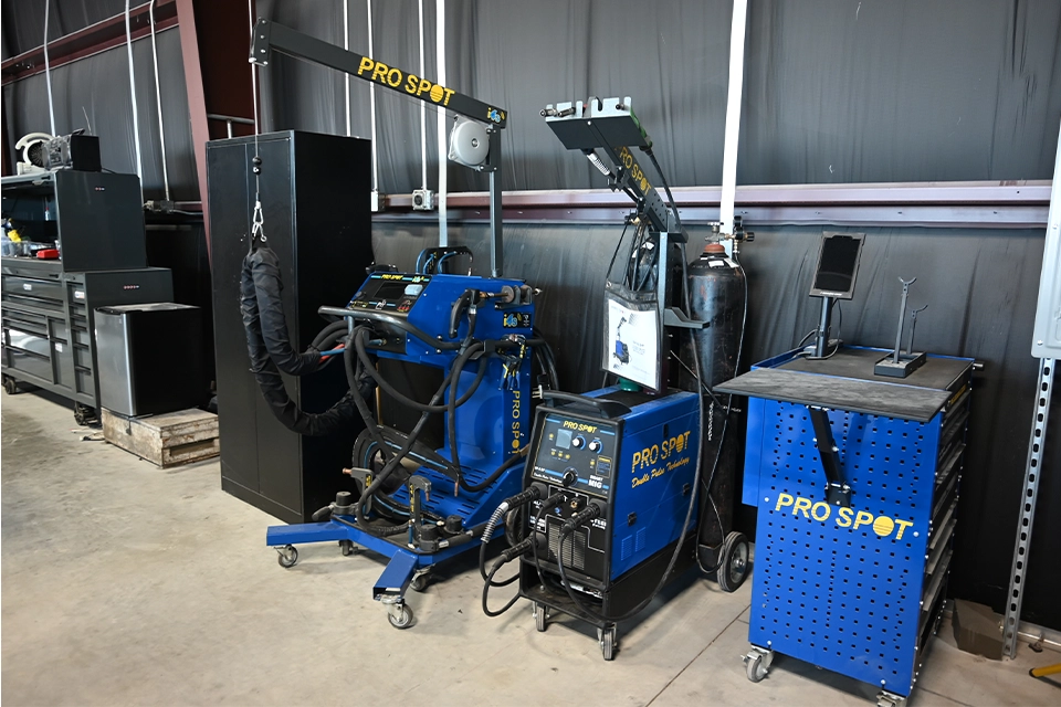 A row of professional auto-body repair equipment inside a workshop, including multiple blue PRO SPOT welding and dent-repair machines, a tall black storage cabinet, tool chests, and a wheeled work table. The machines have cables, hoses, and articulating arms attached, and the workspace is set against a dark metal wall in an industrial shop setting.